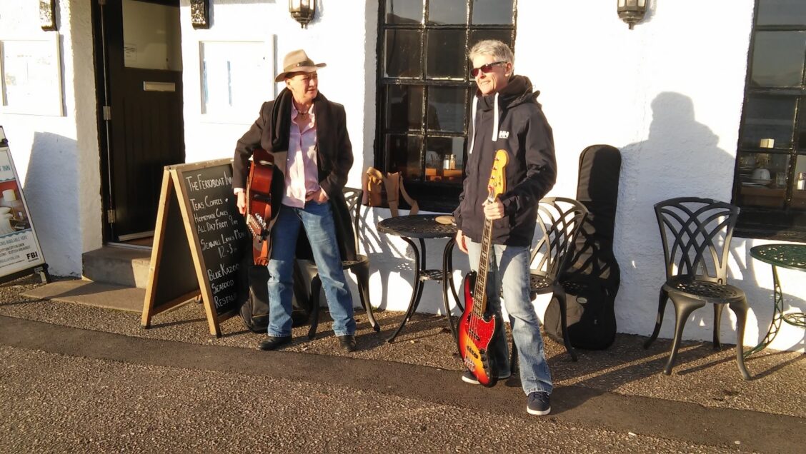 Two women with short hair and guitars smile outside a pub
